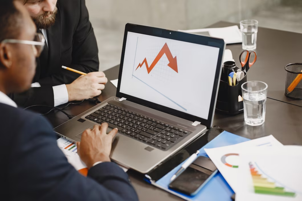 Two men looking at a laptop screen with a downward arrow on an office table.