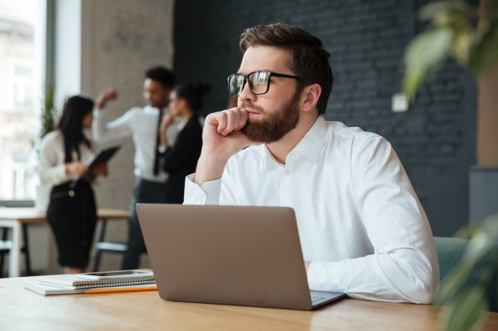 A man with eyeglasses, sitting in front of a laptop, resting his hand on his chin.