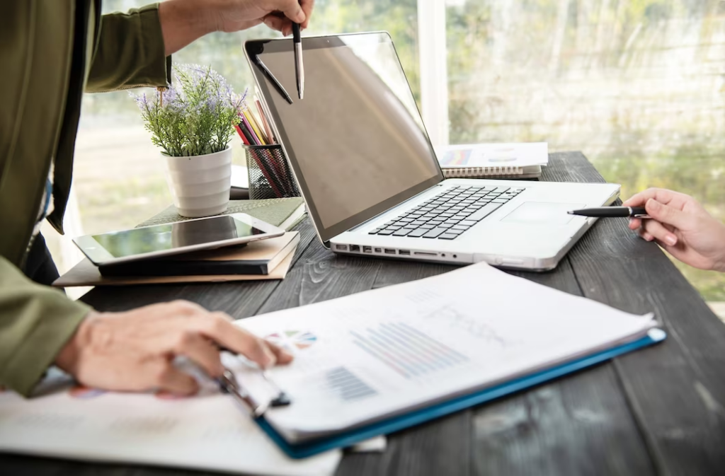 A hand holding papers and pointing at something on a laptop