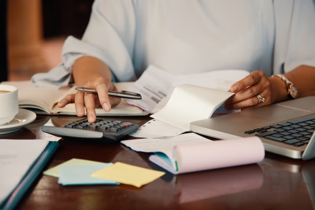 Hand holding a pen and typing on a calculator, while the other hand holds a paper